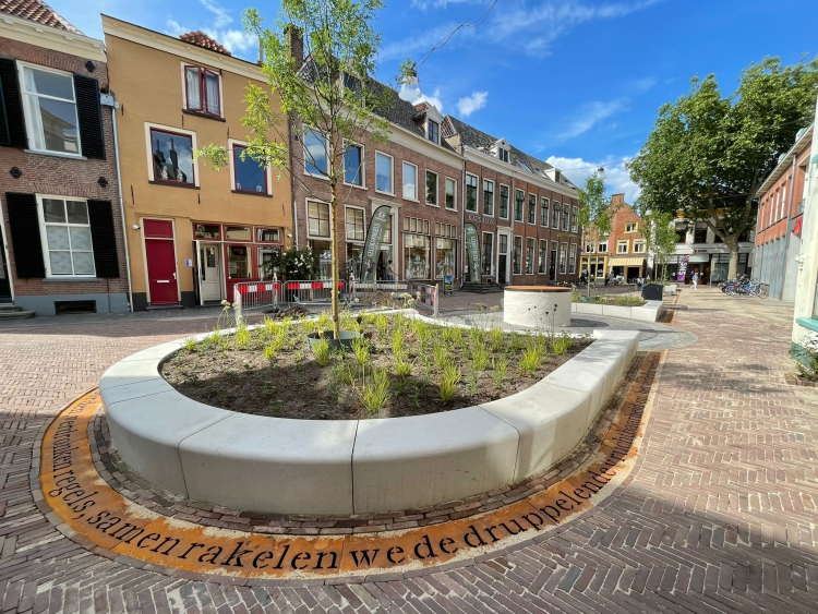 Het heringerichte plein met vooraan een plantenbak met boom en een regenwatergeul met gedicht.